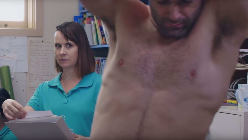 A woman in a turquoise polo neck looks at a topless man in an office.