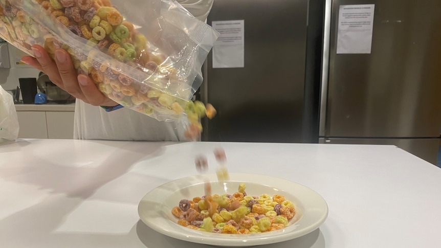 Fruit loops are being poured out of a see through packet into a white bowl.