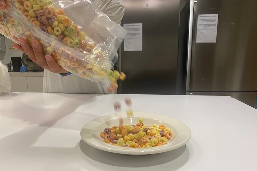 Fruit loops are being poured out of a see through packet into a white bowl.