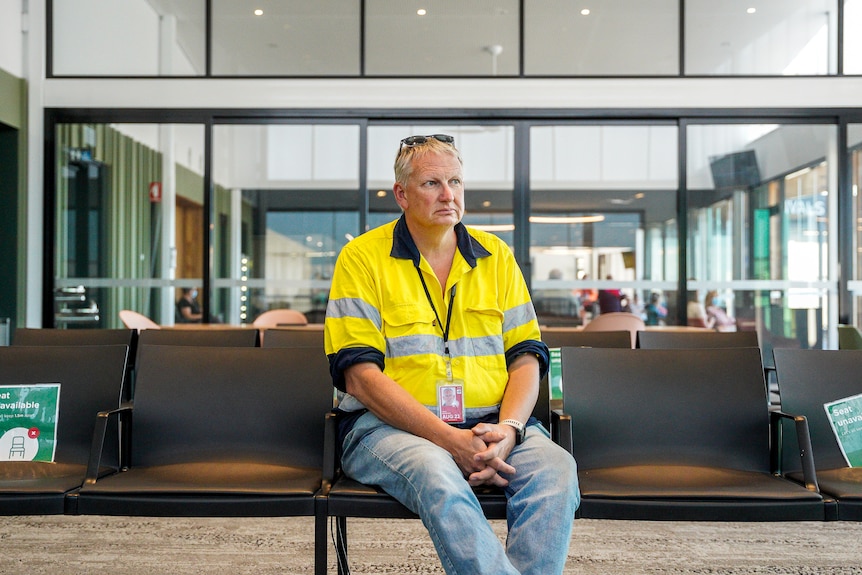 A high visibility man sits in an airport terminal chair and looks out over the tarmac.