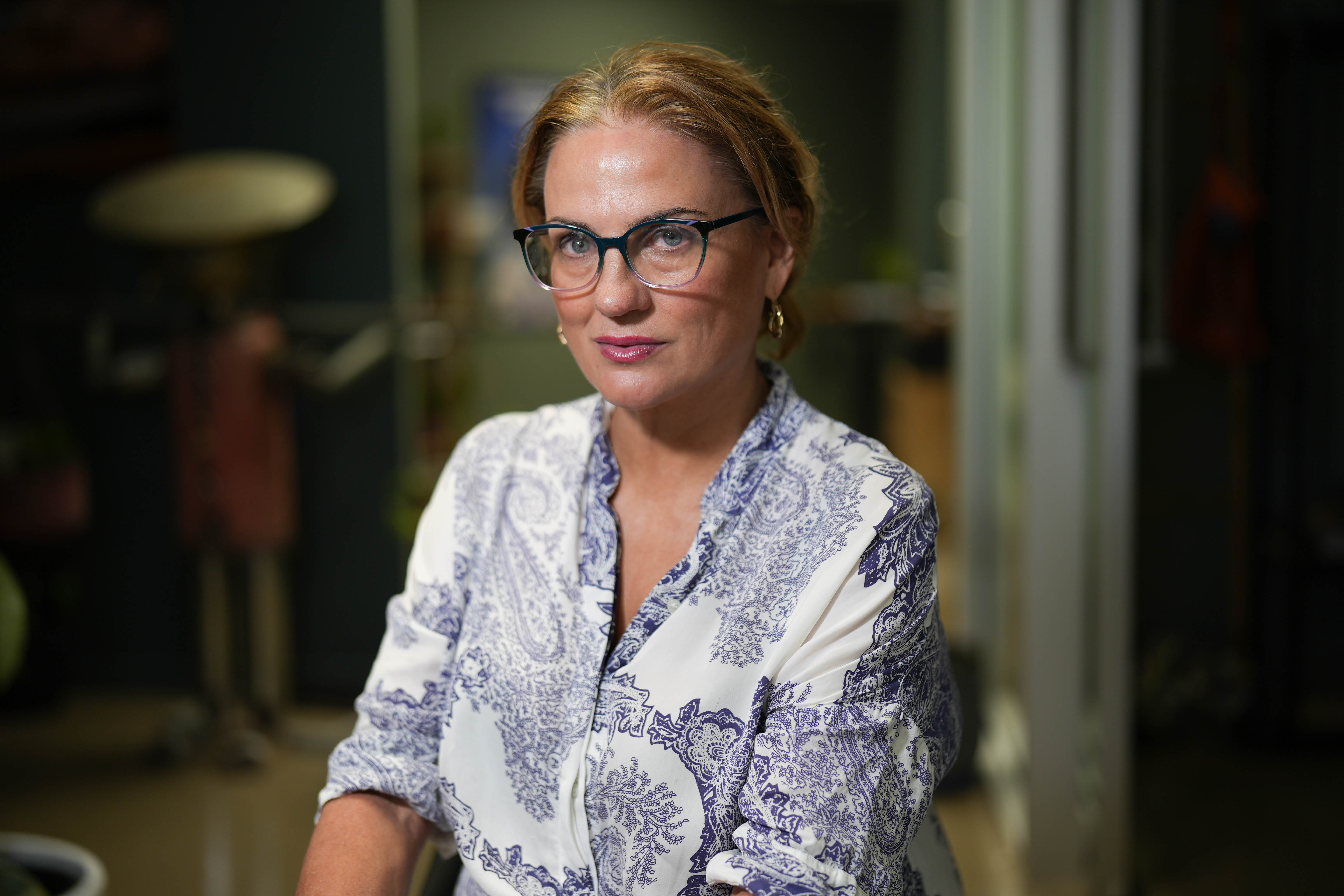 A woman with glasses looks at the camera with her hair tied up and a blue and white t-shirt. 