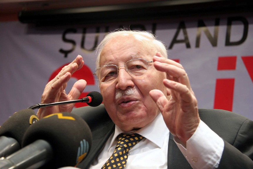 Former Turkish prime minister Necmettin Erbakan speaks into microphones while gesturing with his hands.