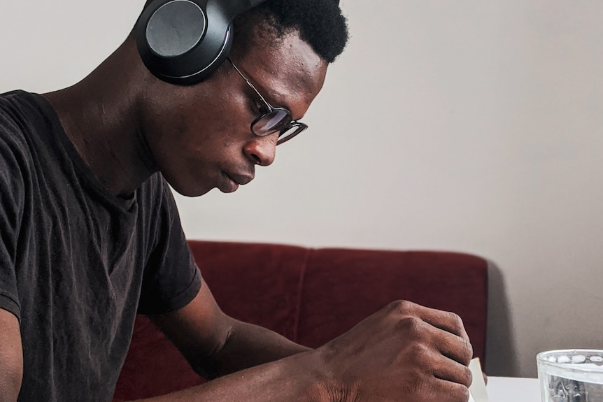 A black man wearing noise-cancelling headphones and reading glasses, bent over a book he is reading.