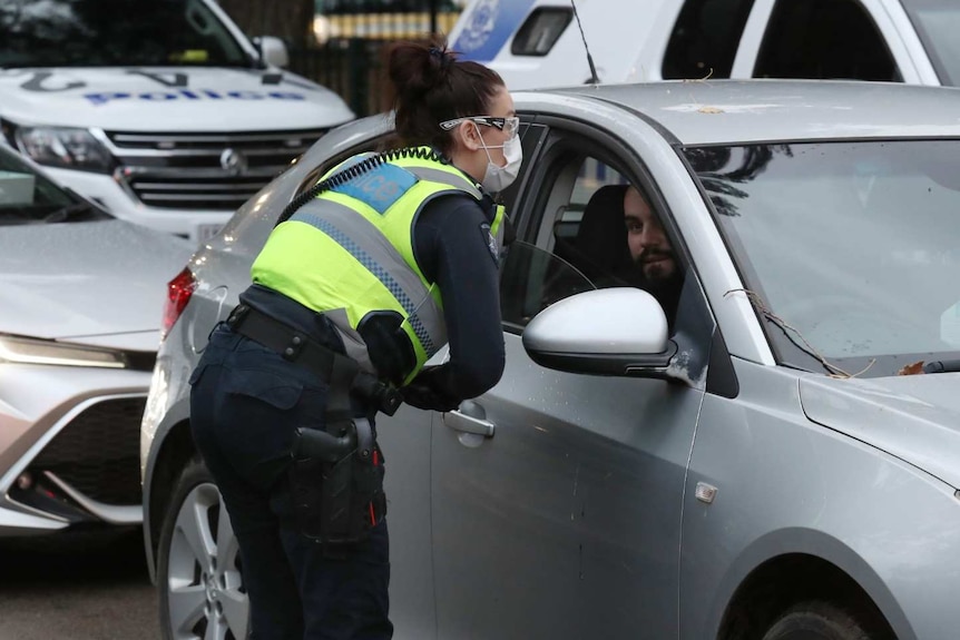 A female police officer speaks to a man through his car window. Police cars can be seen in the background.