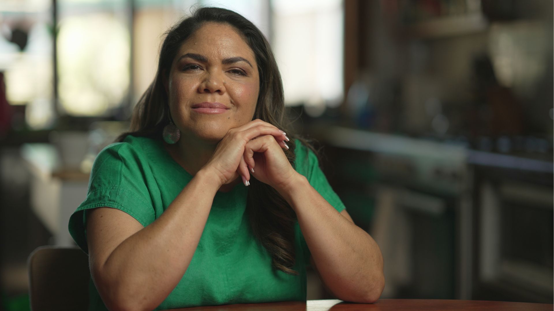 Woman wearing green top holds her hands together under her chin while seated at a dining table