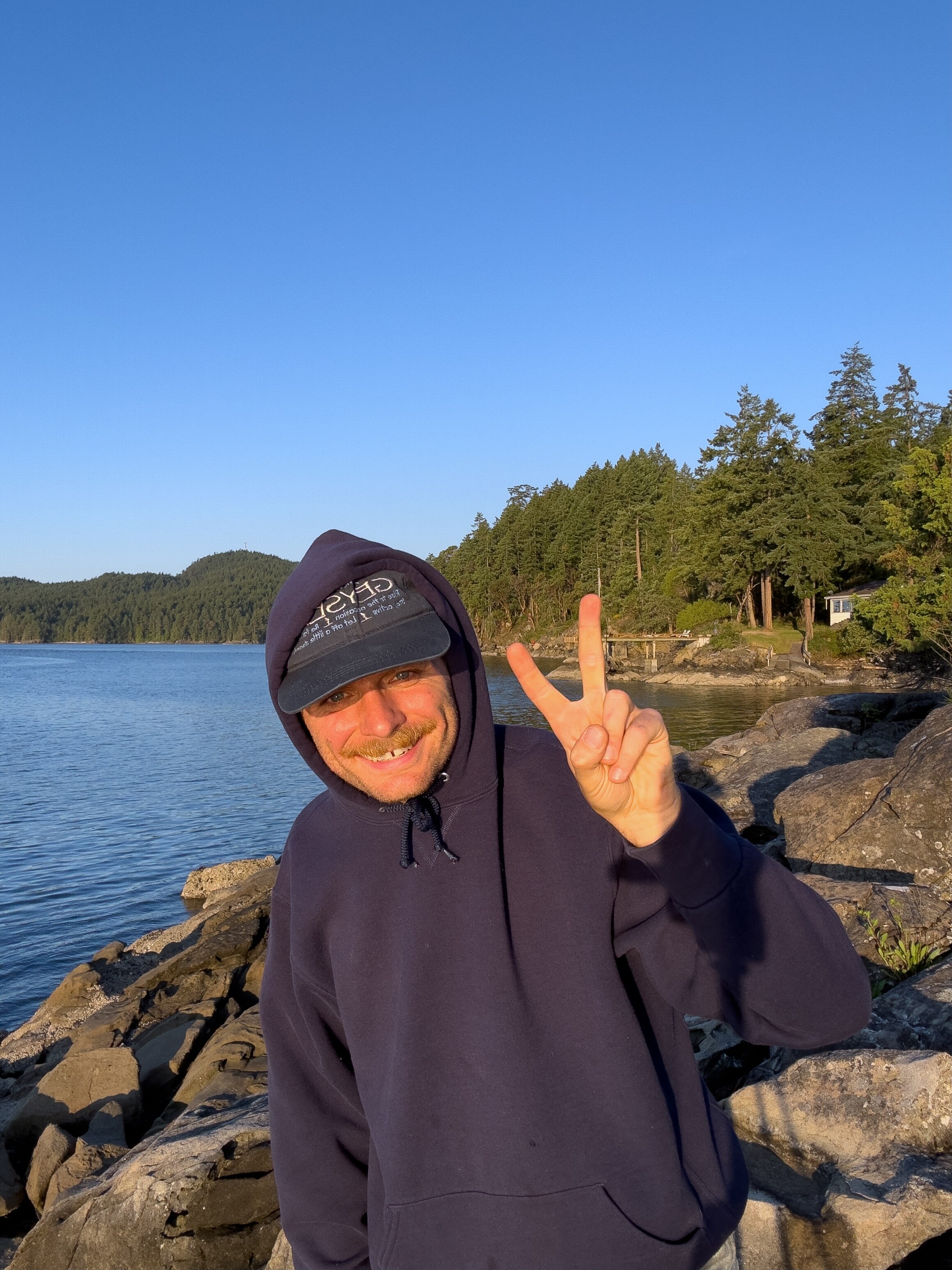 Mac DeMarco does a peace sign standing on rocks on the edge of a lake with a green forest and water behind him