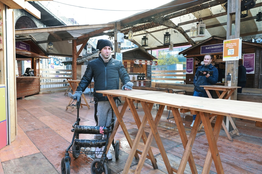 A man in a puffer jacket and beanie standing with a walker at a trestle table at a bar
