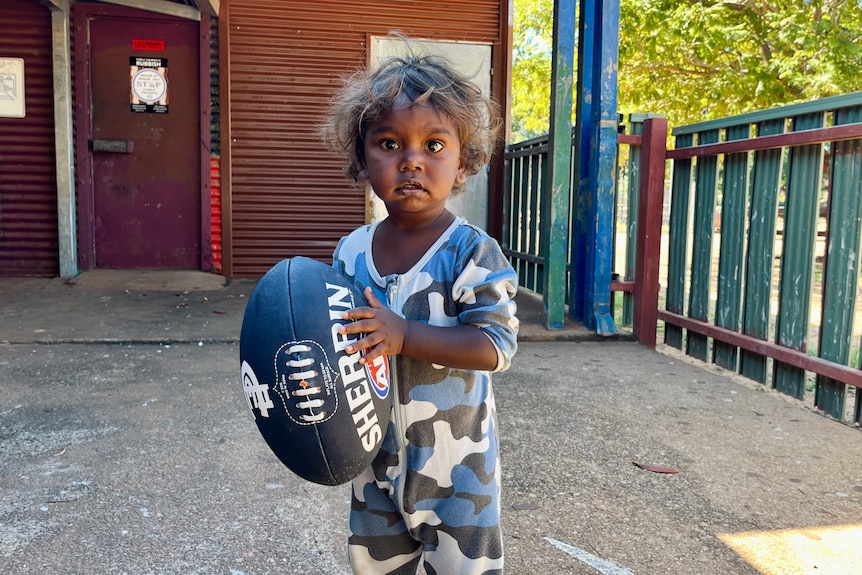 A small child stands outside holding a football