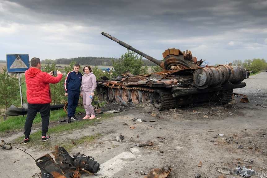 A man takes a photo as a man and woman pose in front of a destroyed tank in a field