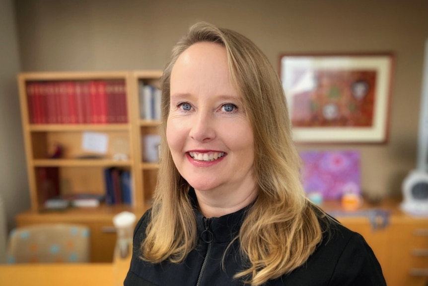 A woman smiles at the camera in an office with a painting and books in the background.