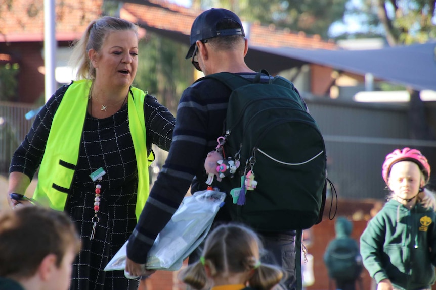 A teacher with a bright yellow vest talks to a parent standing with children wearing school bags and uniforms.