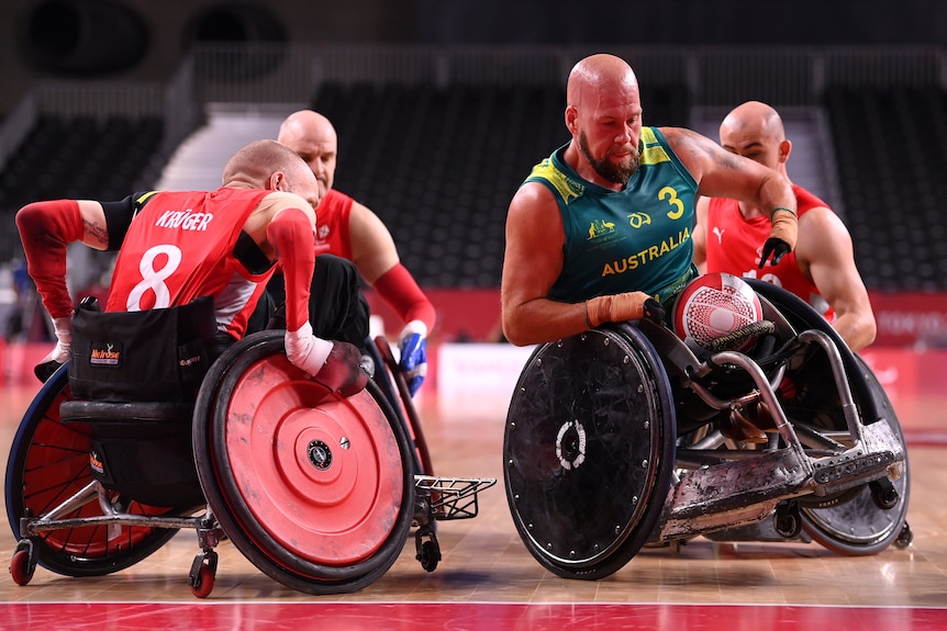 Wheelchair rugby player in green and gold drives with the ball with defenders around him.