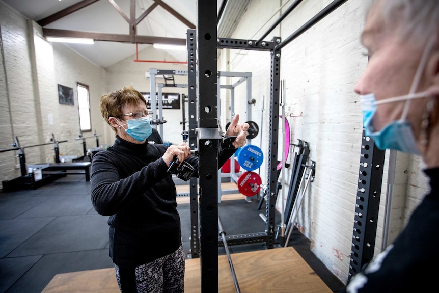 A woman sets up a weight machine while another looks on. Both are wearing covid masks