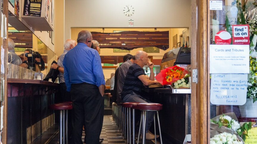 Customers sit on bar stools drinking their coffees as men embrace behind them.
