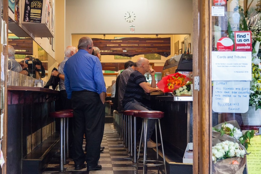 Customers sit on bar stools drinking their coffees as men embrace behind them.