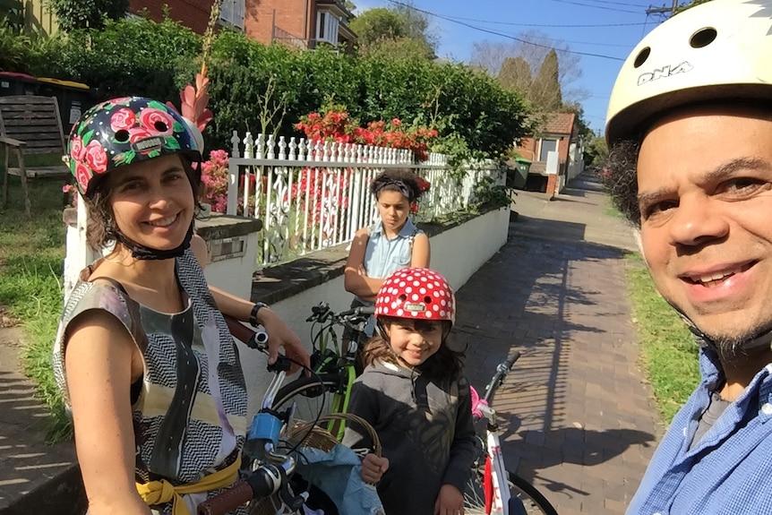 A woman, man and two girls stand next to bicycles on a sunny suburban footpath in a selfie-style photograph