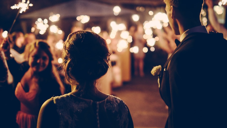 A silhouetted couple stand before a crowd of people with sparklers being waved in a blurry scene.
