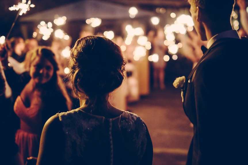 A silhouetted couple stand before a crowd of people with sparklers being waved in a blurry scene.