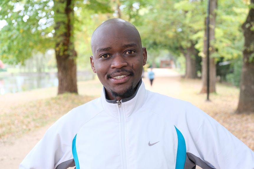 A close portrait of a young man wearing a white jacket looking ahead with a park in the background.