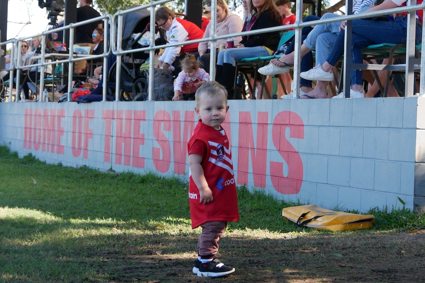 A young boy in a red and white AFL uniform in front of a grand stand with the word ""Home of the Swans" painted on it