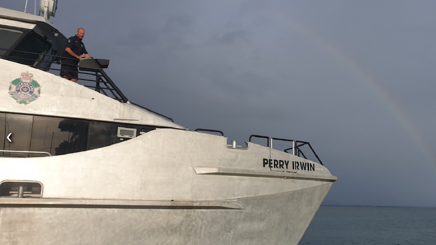 A policeman stands on the deck of a police boat, a rainbow visible on the horizon