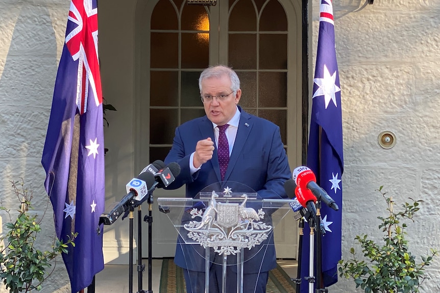 Scott Morrison standing at a podium in front of two australian flags in front of a sandstone house