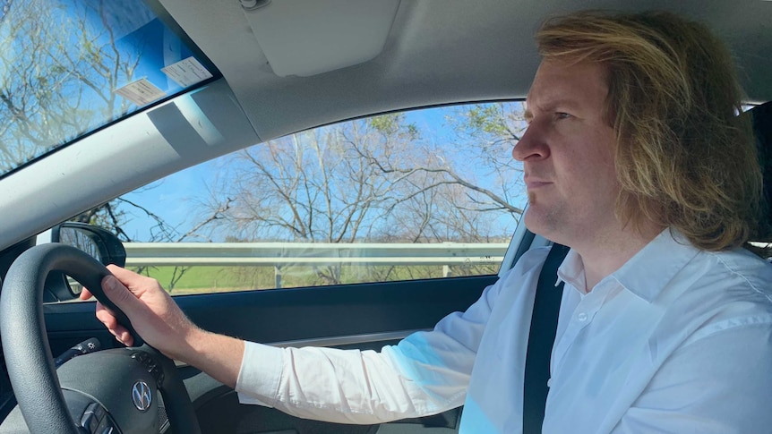 A man driving a car. He's wearing a white button up shirt and has chin-length blonde hair.