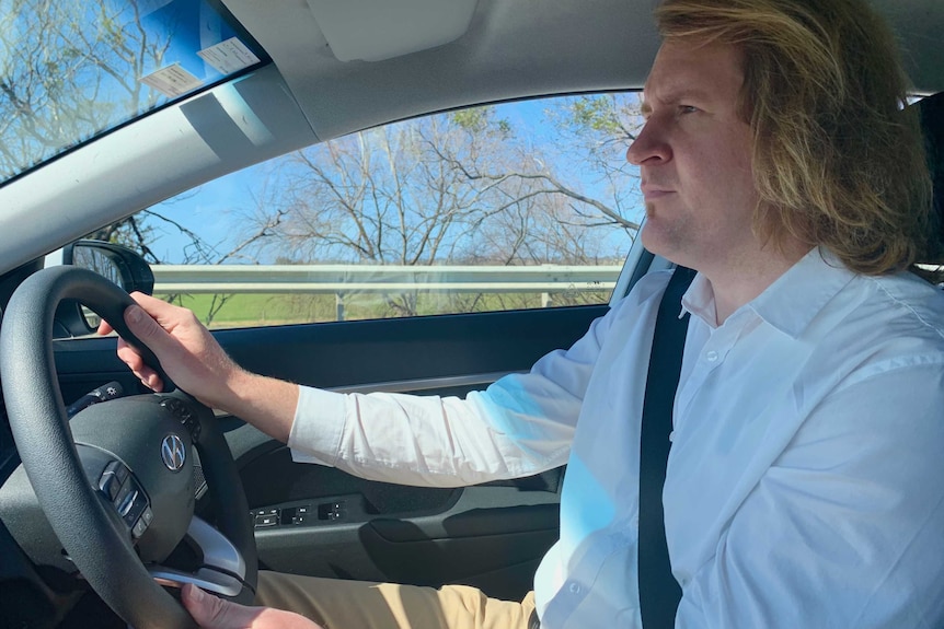 A man driving a car. He's wearing a white button up shirt and has chin-length blonde hair.