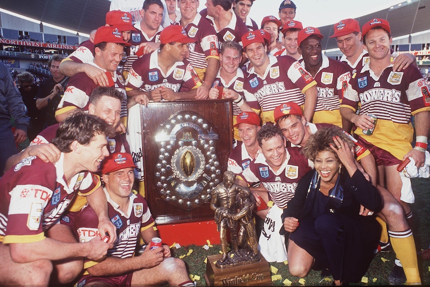 Tina Turner, laughing, crouches next to a team of male rugby players surrounding the NRL championship shield.