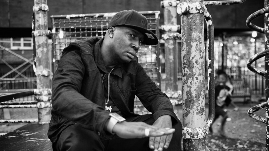 Black and white photo of hip hop icon Grandmaster Flash sitting on a street corner wearing a baseball cap and looking stern