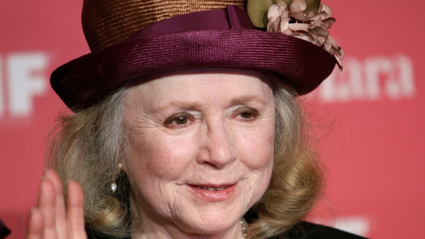 Piper Laurie wearing a purple hat on a red carpet holds up her right hand to wave