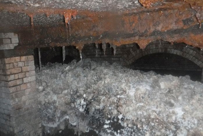 A grey and white mass of congealed fats and household items blocks two thirds of an old brick sewer.