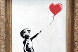 A black and white painting in a thick gold frame of a girl reaching into the air as a love-heart shaped balloon floats away