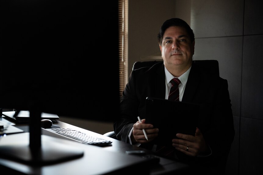 A man in a black suit in a dim, naturally lit room with a computer and table.