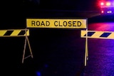 A black and yellow road closed sign illuminated by police lights at night.