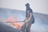 Firefighter wearing a mask and overalls with a fire burning on a hill behind him