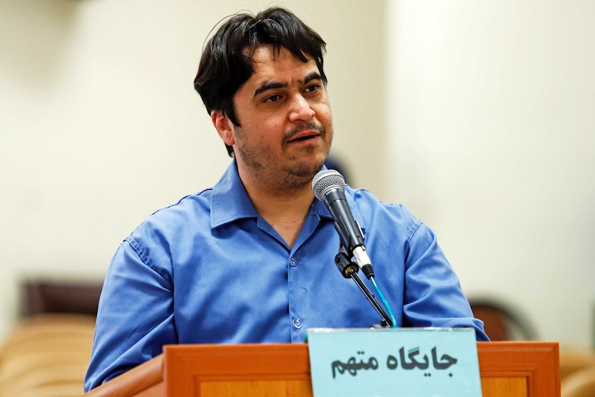A man with dark hair an blue button-up shirt stands speaking in front of a microphone.