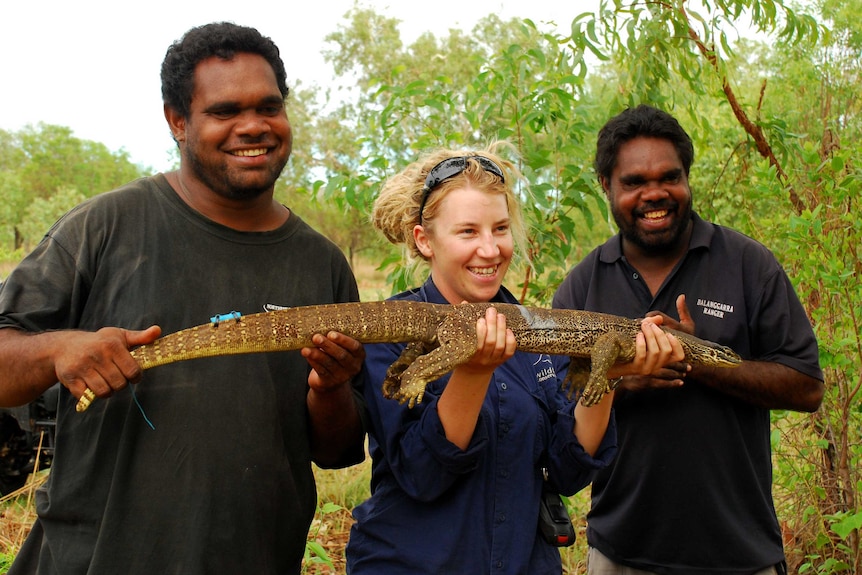Three people hold up a goanna smiling