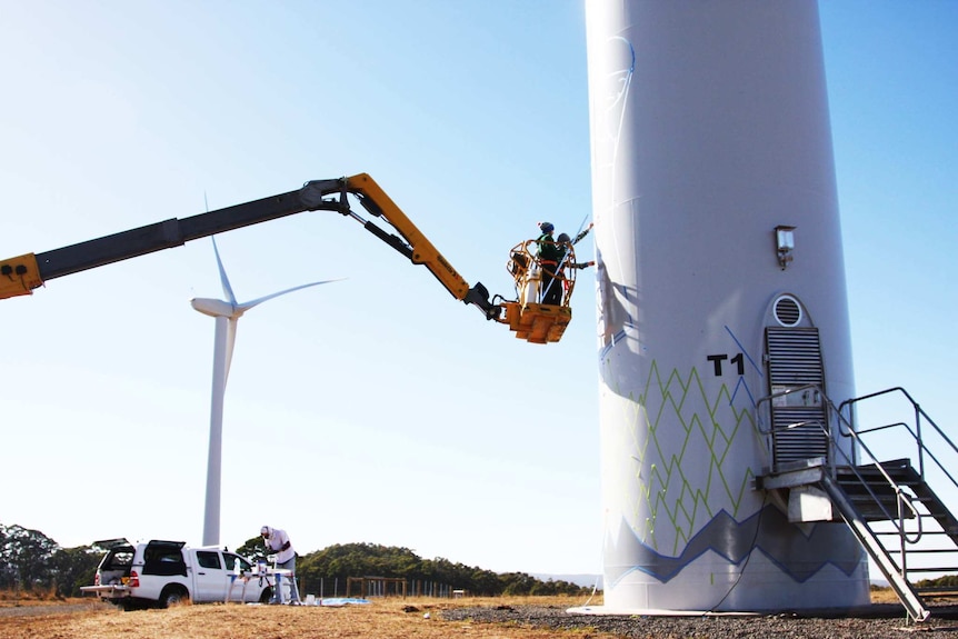 A cherry picker holds a man up to a wind turbine so he can paint it.