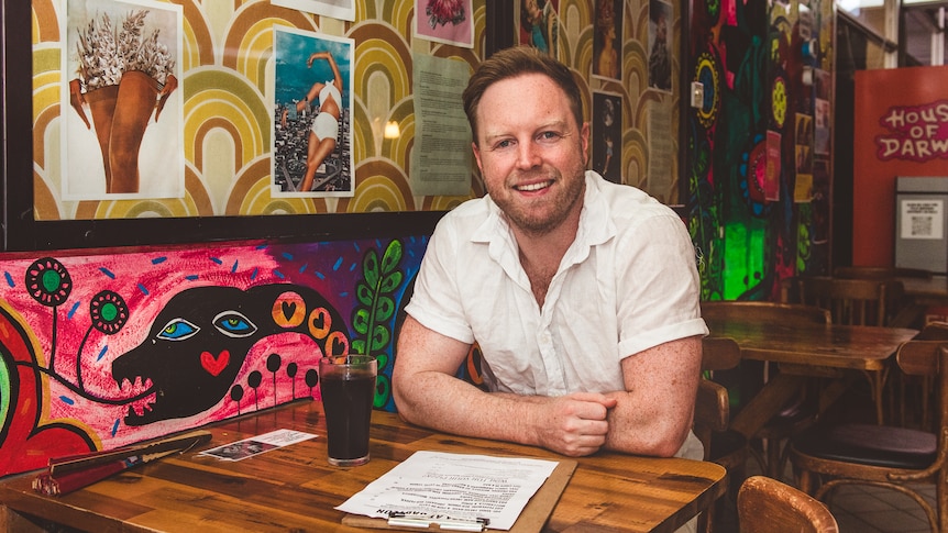 James Findlay sitting at a cafe table with a menu. The wall behind him is covered in funky wallpaper and posters.