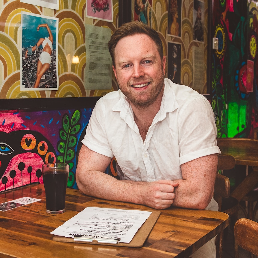James Findlay sitting at a cafe table with a menu. The wall behind him is covered in funky wallpaper and posters.