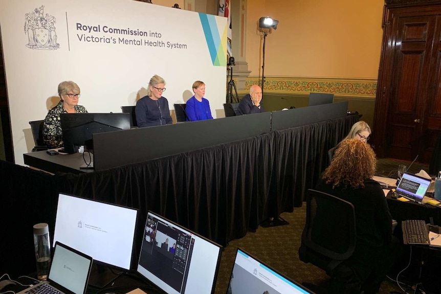 The four royal commissioners sit at a table in a hearing room, while other staff work nearby on computers.