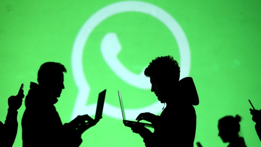 Silhouettes of laptop and mobile device users are seen next to a screen projection of Whatsapp logo