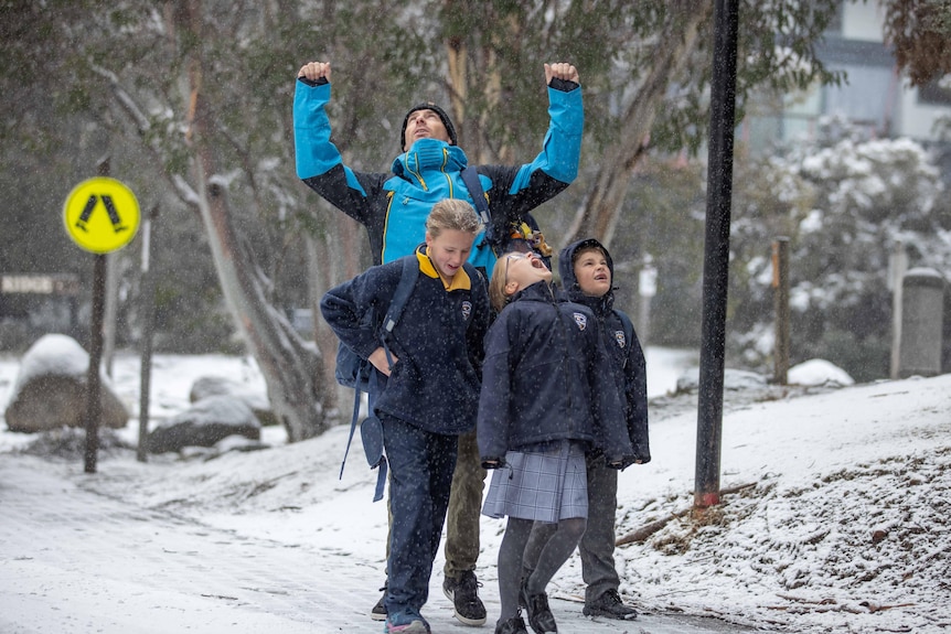 Three children and an adult stand in light snow, one child opens her mouth to catch snowflakes 