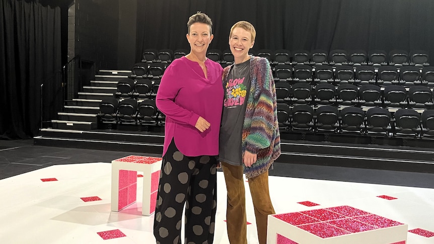 A woman in a pink blouse and a woman with short hair and a eclectic fluffy jacket stand near a pink cube on theatre stage.