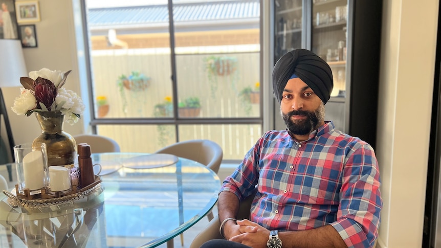 A bearded man in a Sikh turban and checked shirt sits relaxed at a dining table, hands clasped, smiling at the camera 
