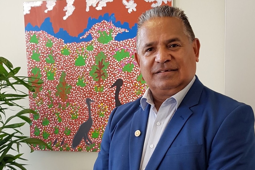 Tony wears a blue blazer pictured in front of an Aboriginal painting