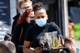 A waitress wears a mask as she carries a tray of alcoholic drinks in an outdoor restaurant as patrons sit on chairs