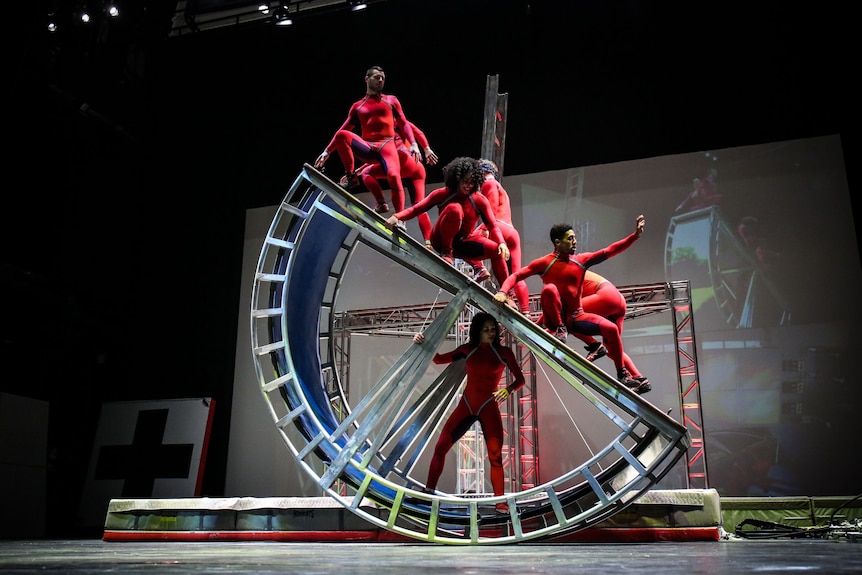 A group of dancers in red unitards straddle a giant prop shaped like a mechanical semi-circle.
