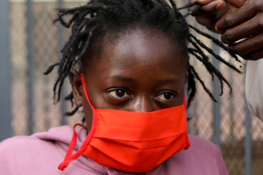 Girl in red mask and pink hoodie gets hair braided while mum in red vest looks on in background